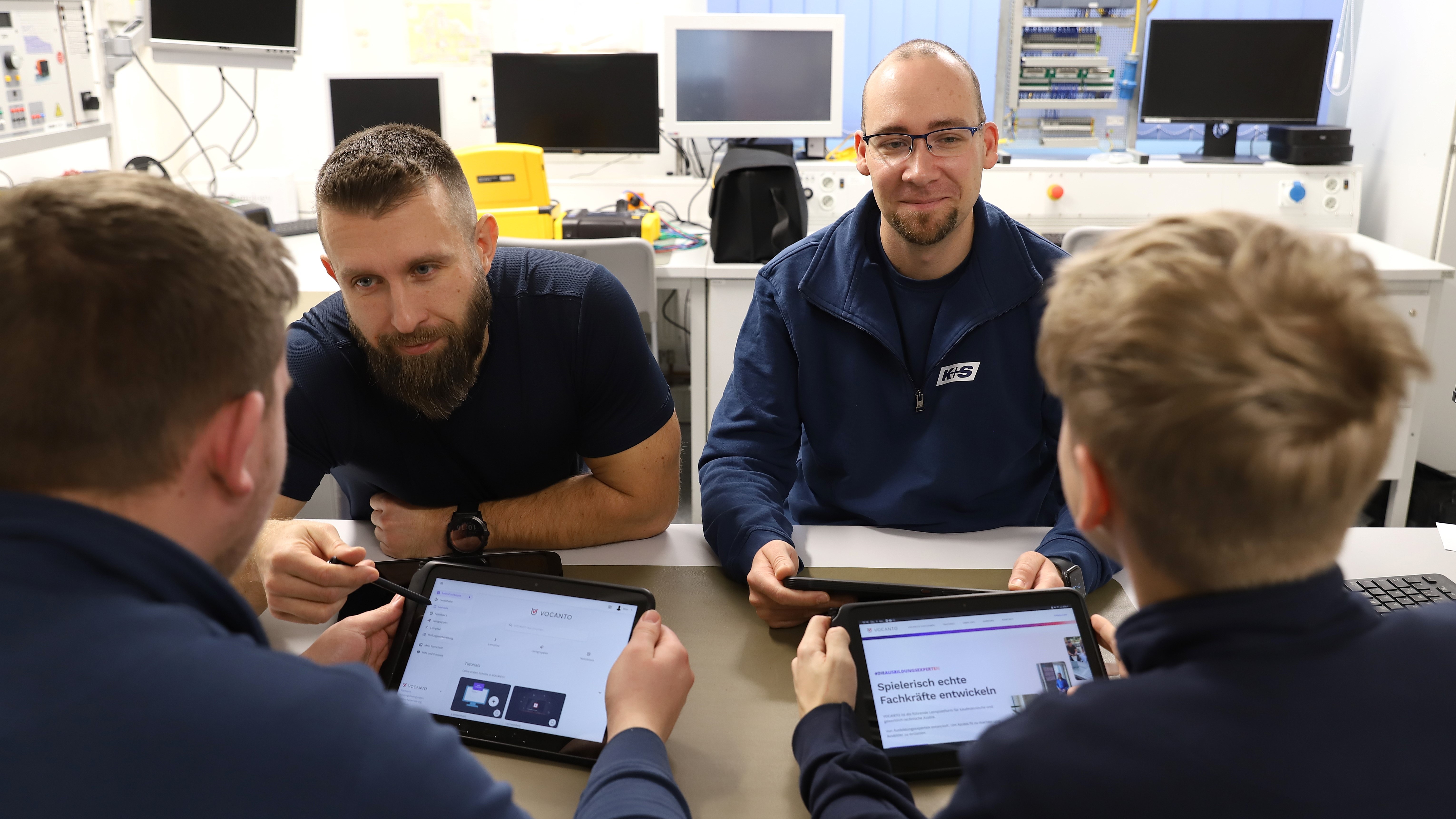 Zwei Auszubildende sitzen ihren Ausbildern gegenüber an einem Tisch und haben ein Tablet in der Hand.